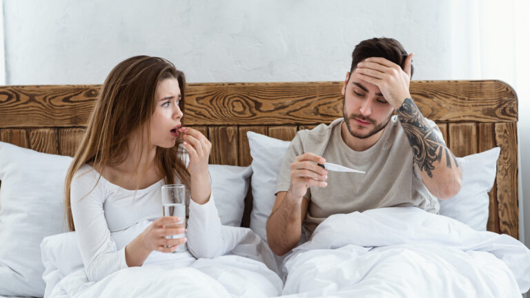 Woman drinking pill, holding glass of water and looking at husband, man touches forehead and looks at thermometer