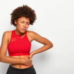 Dissatisfied African American woman holds aching hip, has kidney inflammation, touches location of pain near ribs marked with red dot, wears sport bra, shows perfect figure, has sporty shaped body