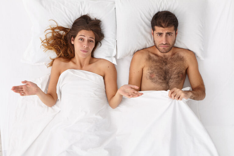 Overhead shot of puzzled woman and her husband have sex problems in bed, displeased expressions, lie under white blanket. Man has impotence, erection failure. Daylife family troubles concept